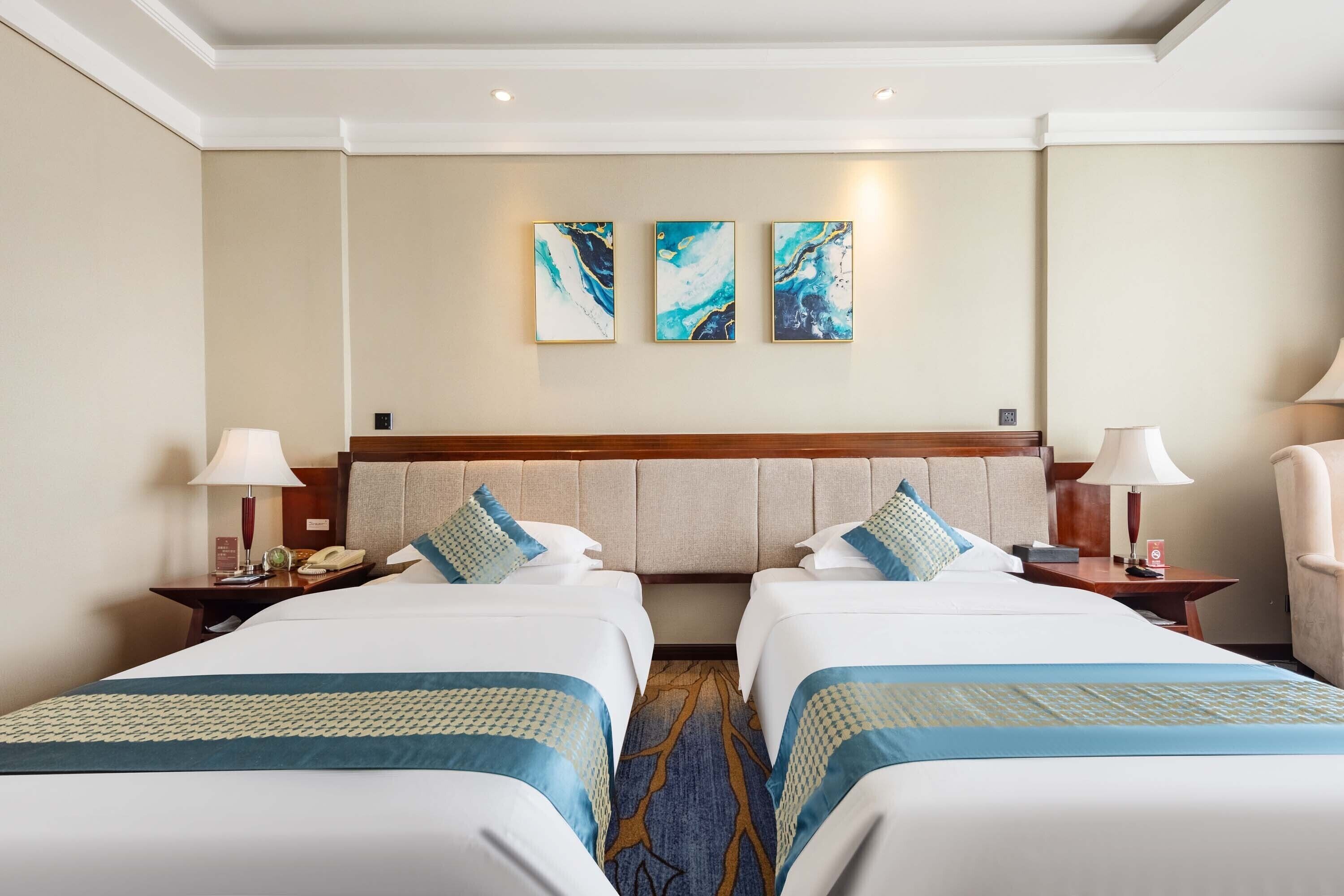 A hotel room with two beds, a headboard, bedside tables, lamps, and artwork on the wall.
