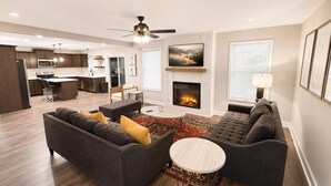 A modern living room with a fireplace, a ceiling fan, and a kitchen in the background.