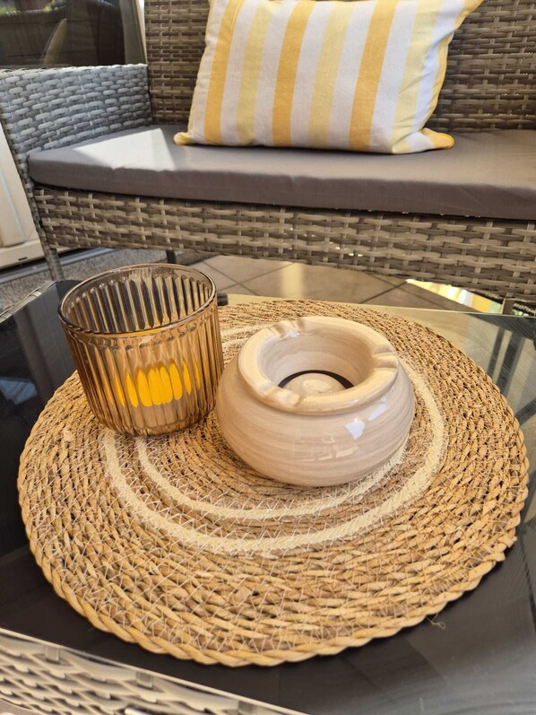 Un canapé en osier agrémenté d’un coussin à rayures, un porte-bougie en verre et un bol en céramique posé sur un set de table en fibres tressées.