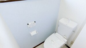 A small, clean bathroom with a toilet, a wall-mounted flush control panel, and a toilet paper holder. - EASE Takasaki Toyooka (Takasaki)