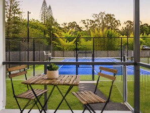 Un balcón con vista a una cancha de tenis, una mesa de madera, dos sillas y una planta en maceta.