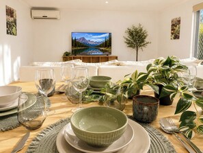 Un comedor con una mesa preparada para cuatro, un televisor mostrando un paisaje de montaña y un sofá al fondo.