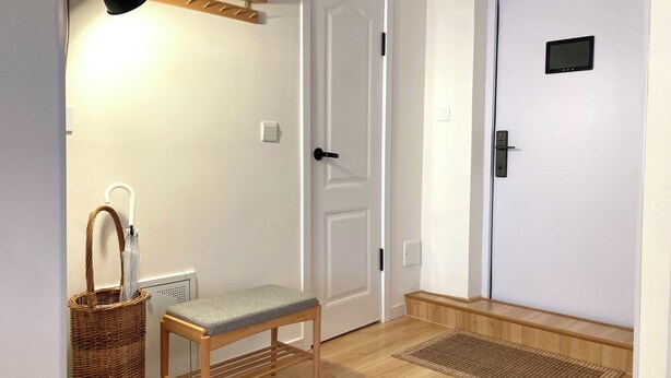 Un pasillo con un banco, una puerta, una lámpara de pared, una cesta y un felpudo.