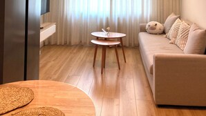 Una sala de estar moderna con una mesa redonda de madera, un sofá con cojines decorativos, una lámpara colgante y un ventanal con cortinas traslúcidas.