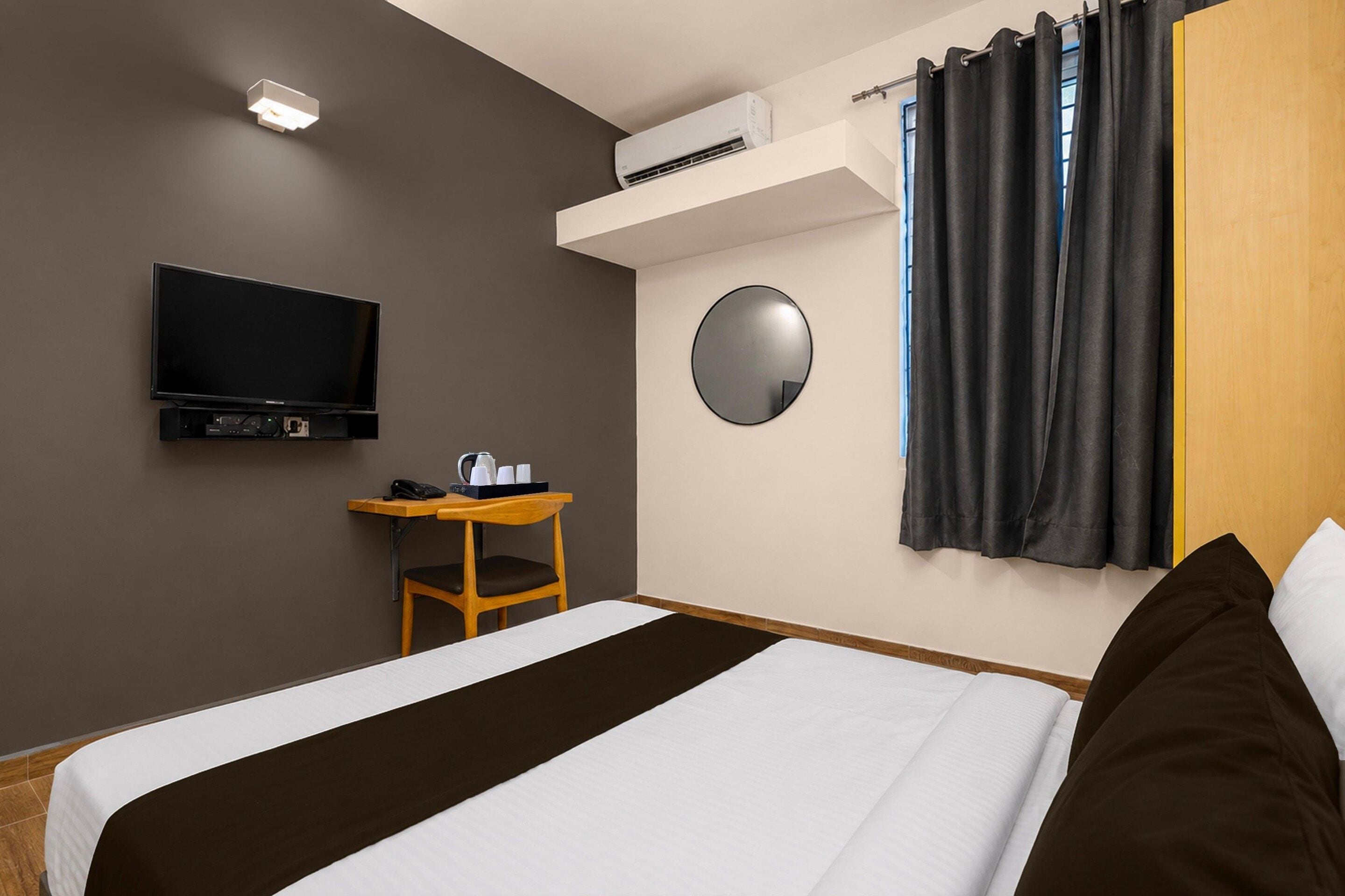 Una camera d'albergo con un letto, una scrivania, un televisore, un condizionatore, uno specchio e una finestra con tende.