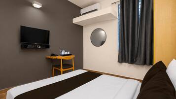 A hotel room with a bed, a desk, a television, an air conditioning unit, a mirror, and a window with curtains.