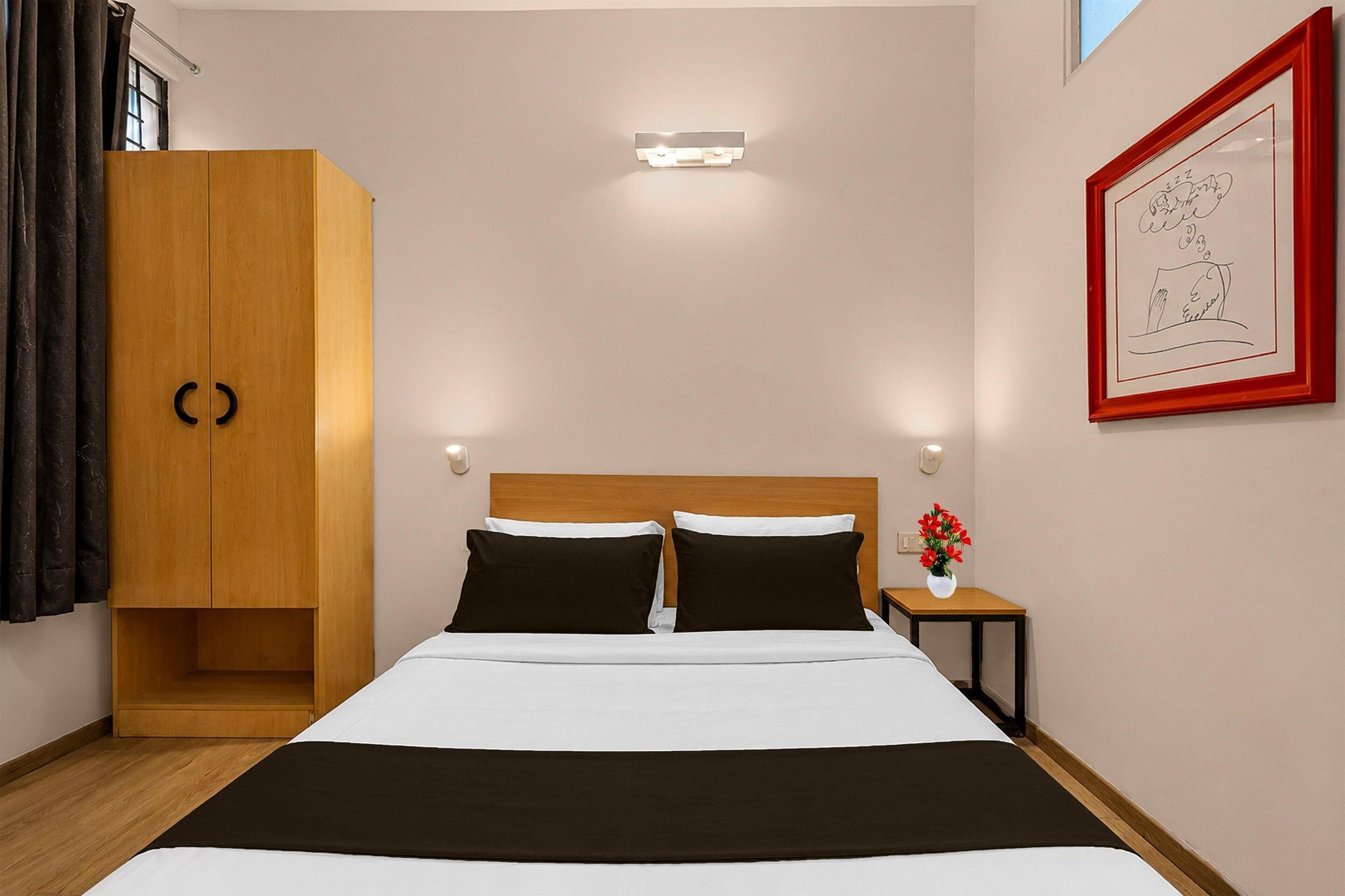 A hotel room with a bed, a wardrobe, a nightstand, a vase with flowers, and a framed picture on the wall.