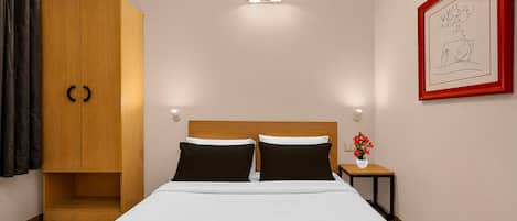 A hotel room with a bed, a wardrobe, a nightstand, a vase with flowers, and a framed picture on the wall.
