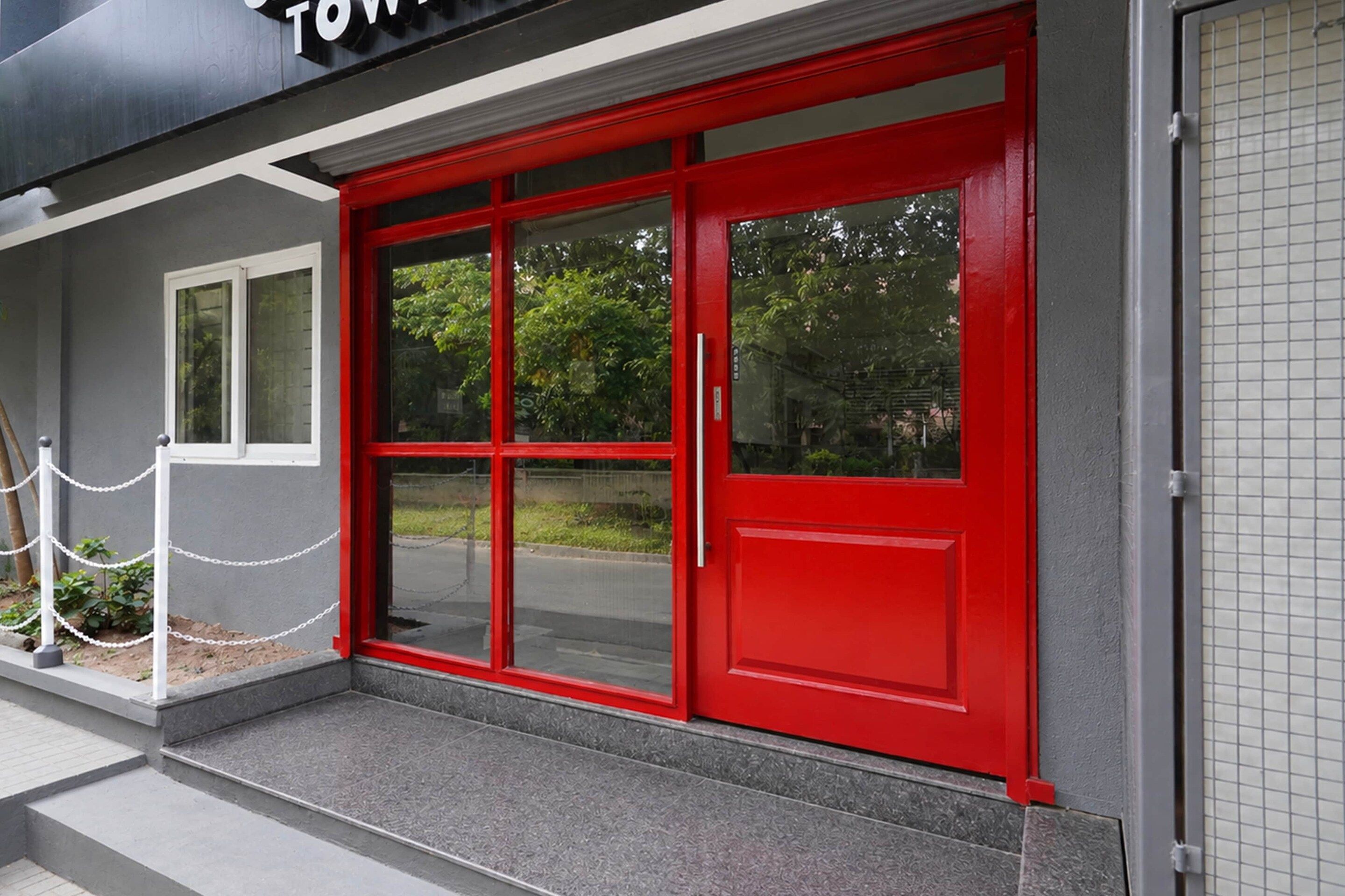 A building entrance with a red door, a white chain barrier, and a grey exterior wall.