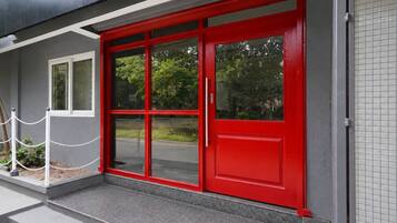 A building entrance with a red door, a white chain barrier, and a grey exterior wall.
