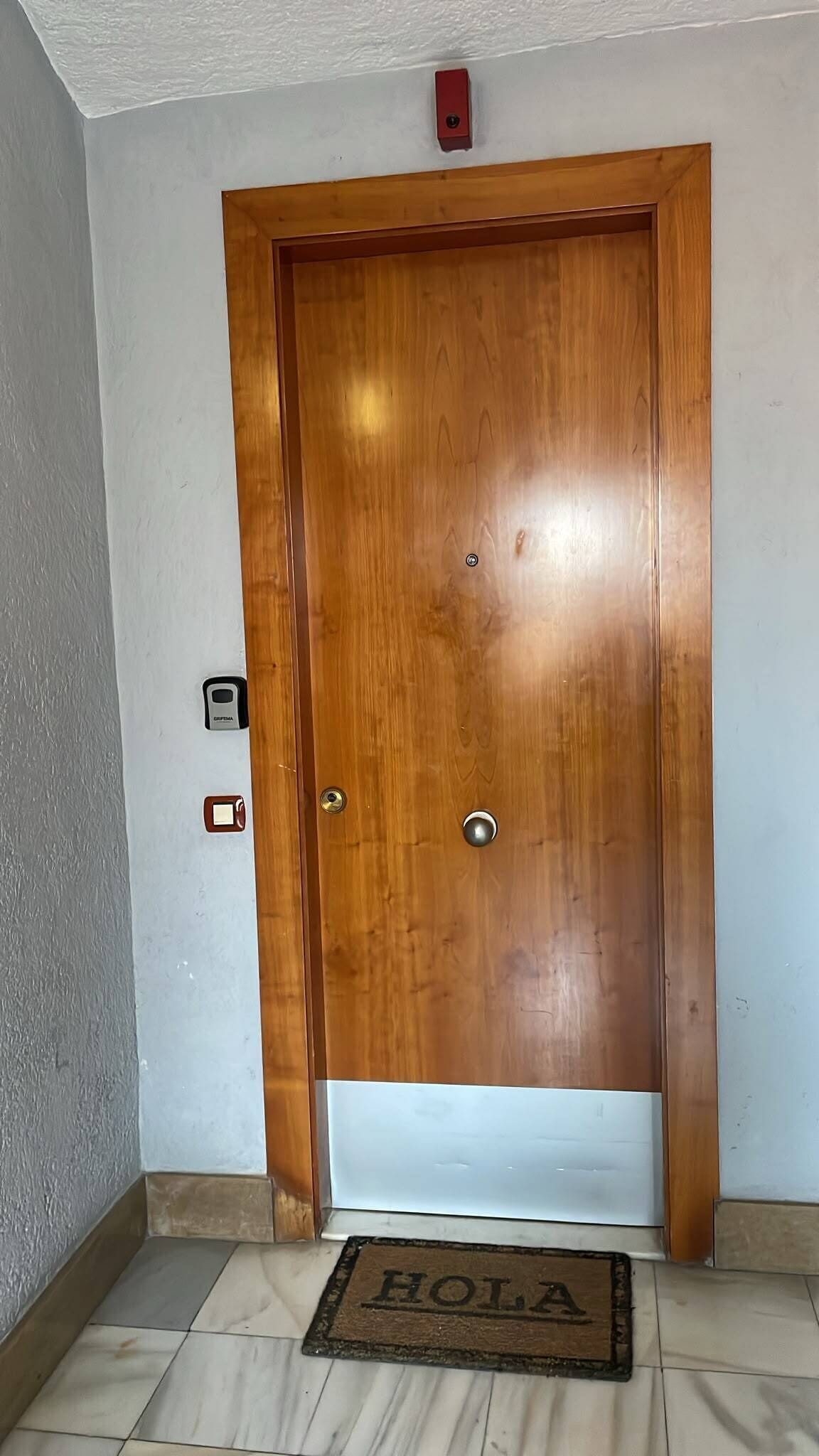 A wooden door with a doorknob and a keyhole, a small intercom, and a doormat with 'HOLLA' on a tiled floor.