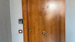 Una puerta de madera con picaporte y cerradura, un pequeño intercomunicador y un felpudo con la palabra 'HOLLA' sobre un suelo de baldosas.
