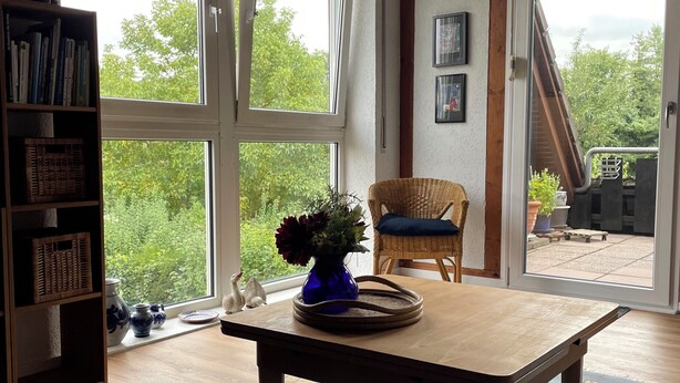 Una habitación con ventanas amplias, una mesa de madera, una silla de mimbre y una estantería.