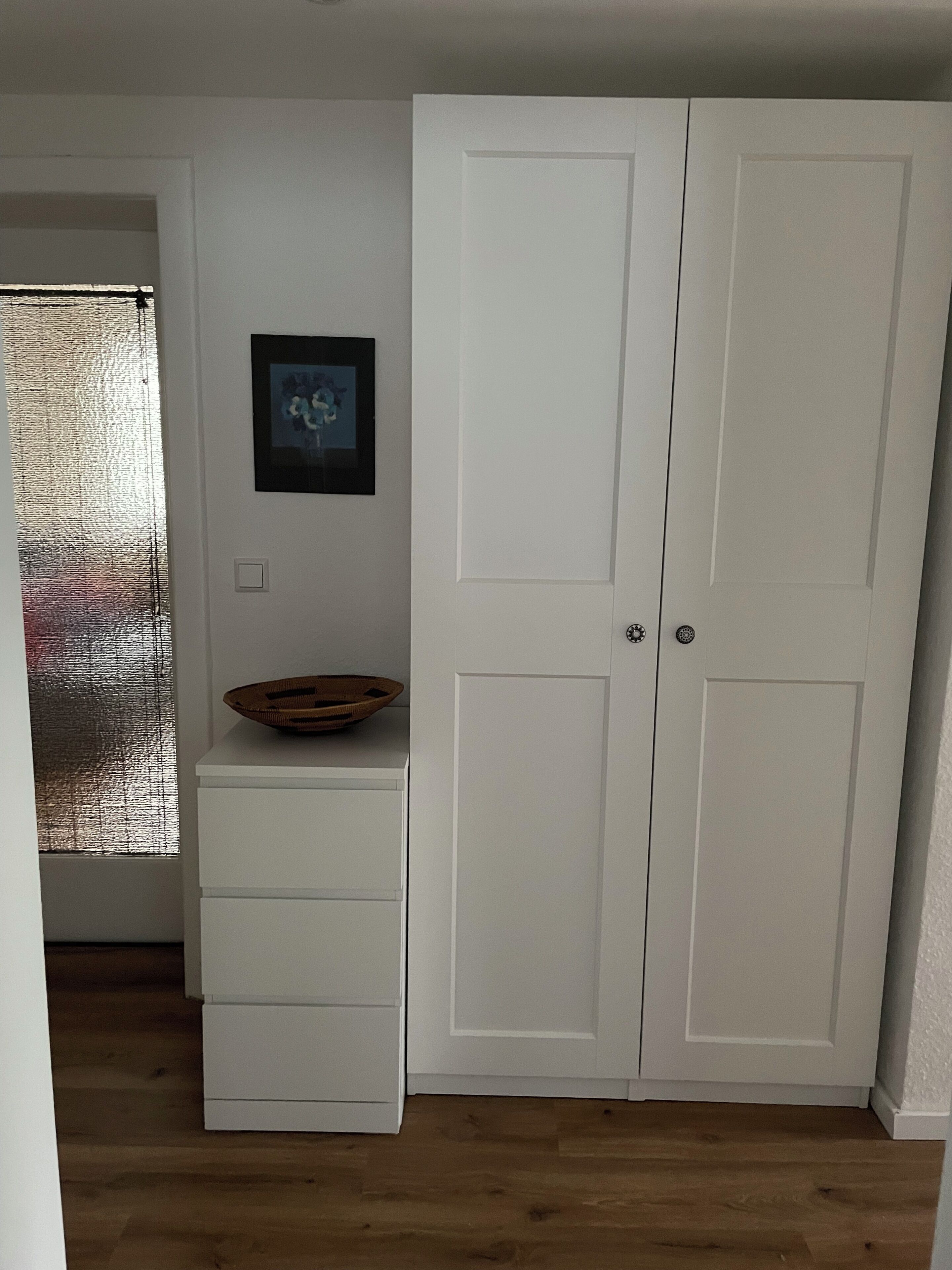 A room with a white wardrobe, a small shelf with a basket, a framed picture on the wall, and a frosted glass window.
