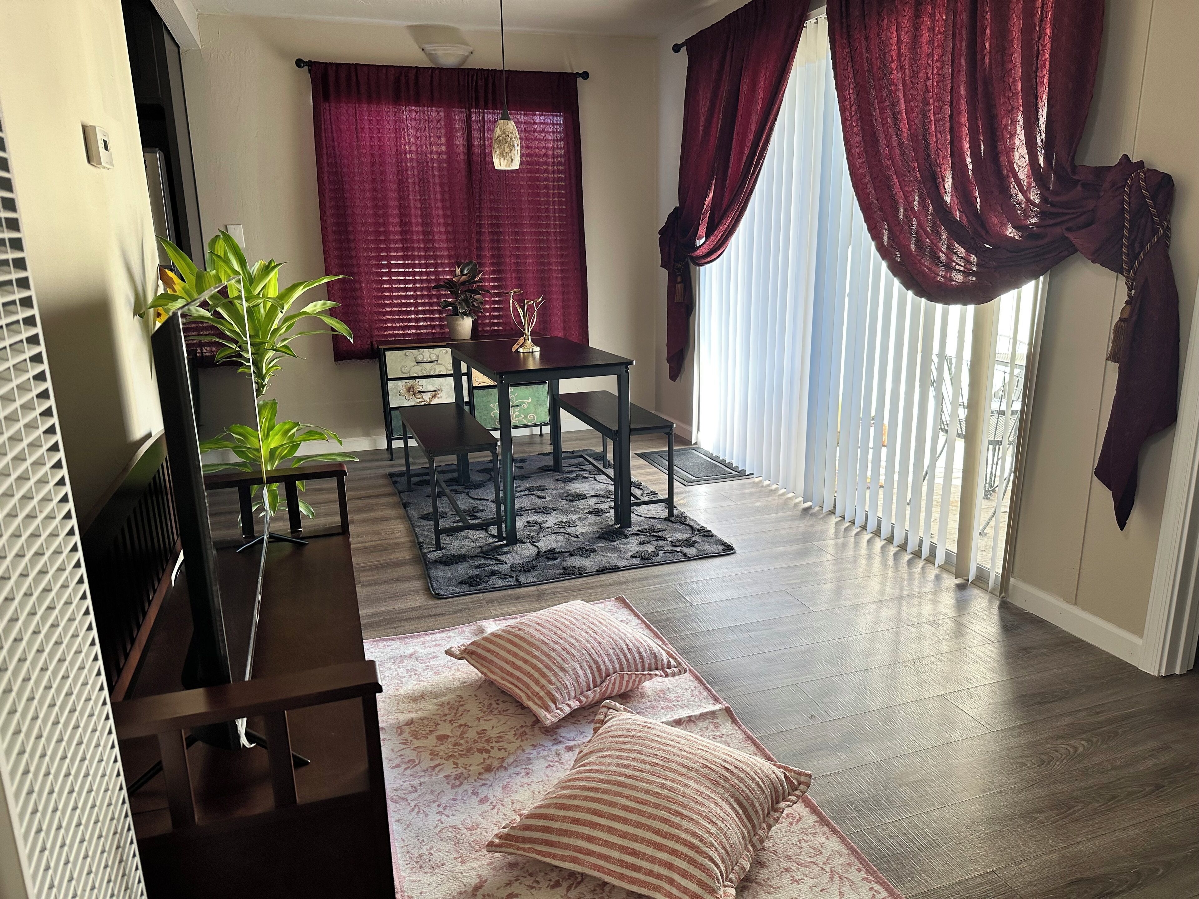 Una habitación con una mesa de comedor, sillas, un banco, un sofá, una planta en maceta y un ventanal con cortinas.