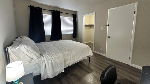 Een slaapkamer met een bed, een bureau met lamp, een stoel, een kast en een deur.
