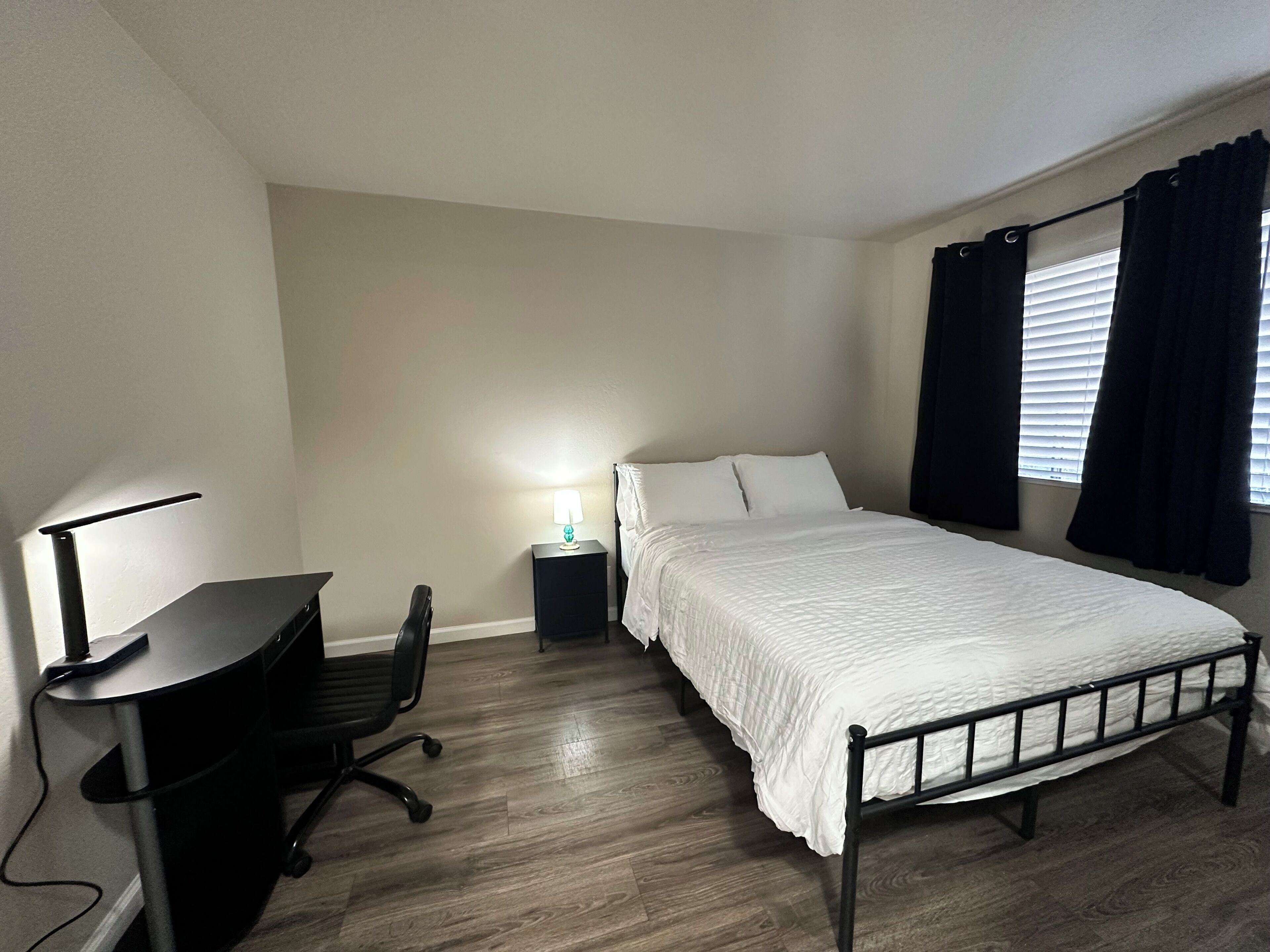 A bedroom with a bed, a desk with a chair, a nightstand with a lamp, and a window with curtains.
