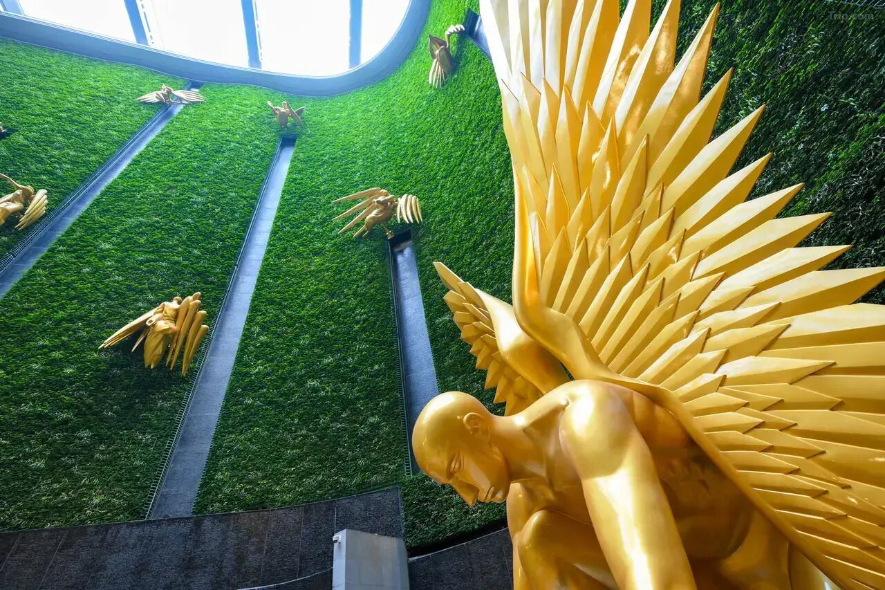 Una estatua dorada de una figura alada, un muro de césped artificial verde y un tragaluz circular.