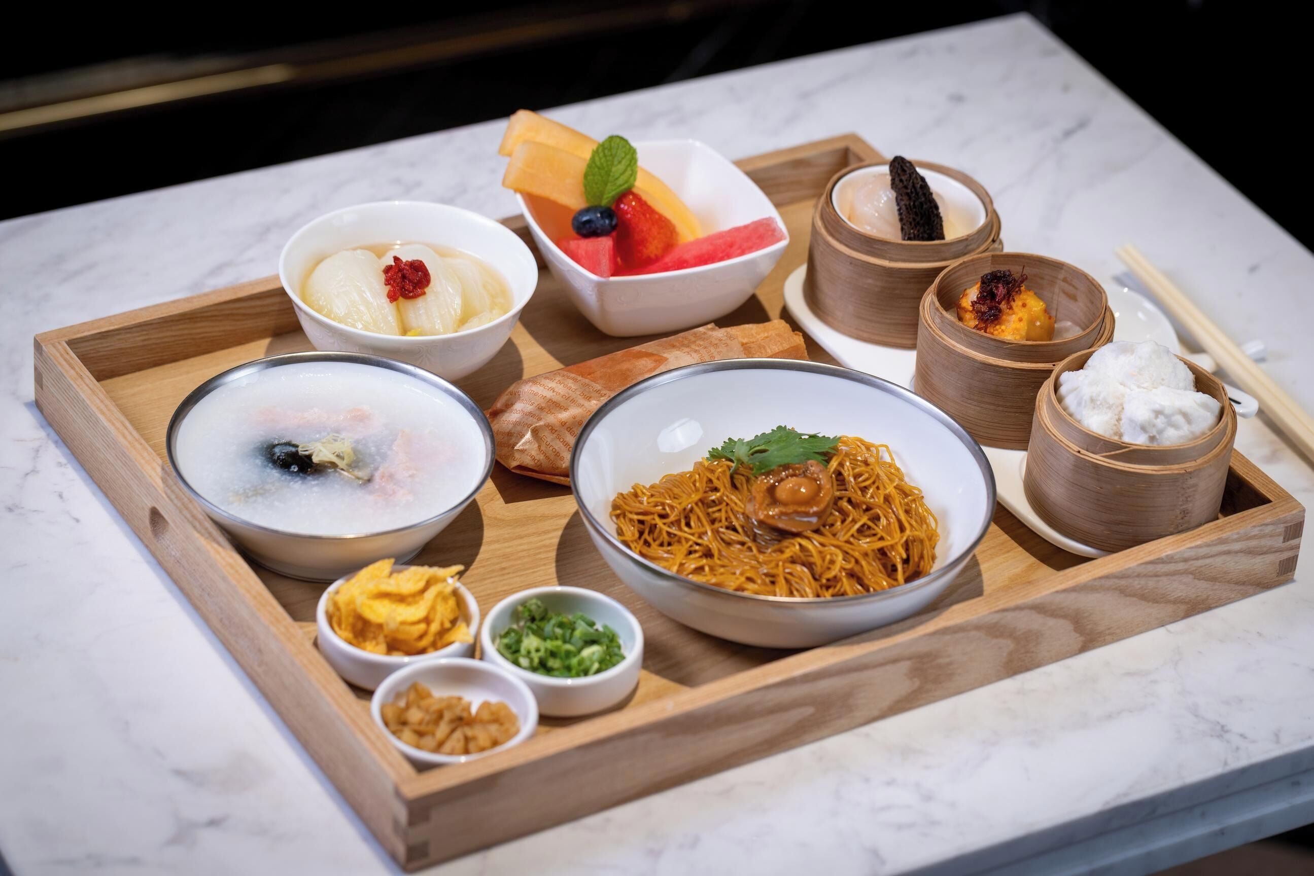 Una bandeja de madera con diversos platos, incluyendo fideos, sopa y fruta.