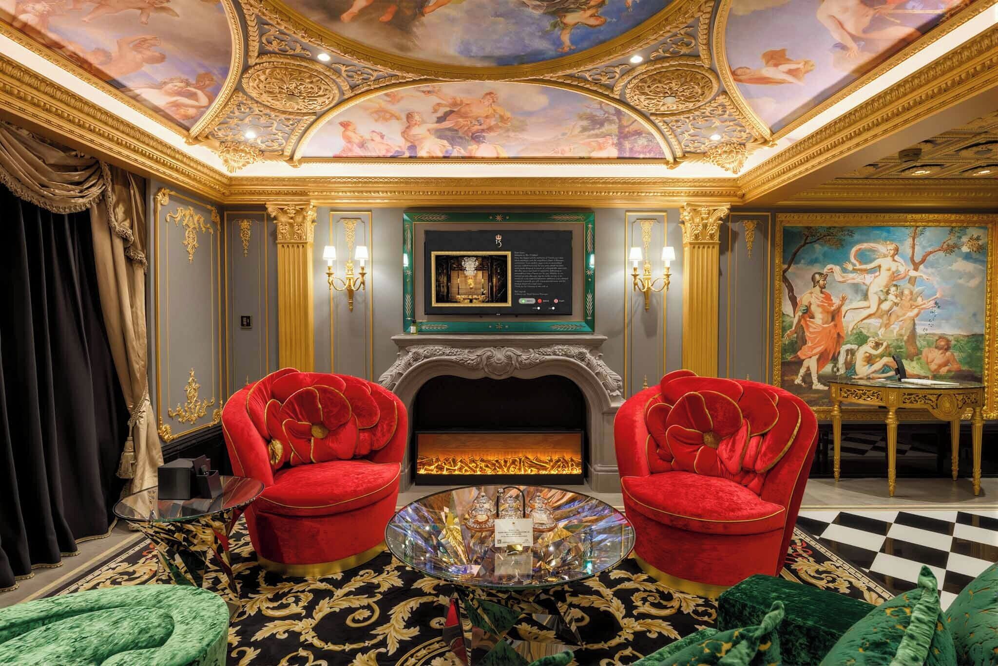 Een luxueuze kamer met een open haard, rode fluweelstoelen, een glazen bijzettafeltje en gedetailleerde kunstwerken aan het plafond.