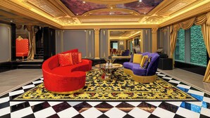 Un hall d’entrée luxueux, orné d’un plafond richement décoré, agrémenté d’un ensemble de canapés rouge et violet, et recouvert d’un tapis à motifs.