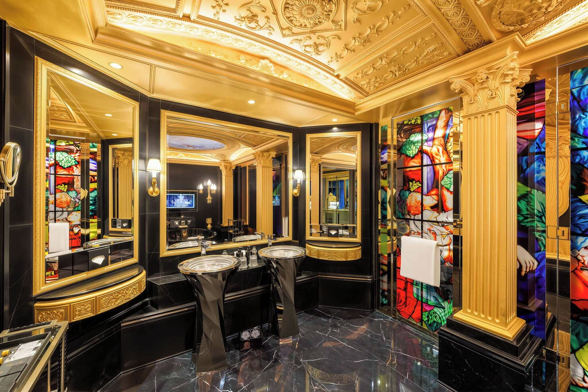 A luxurious bathroom with ornate gold detailing, multiple mirrors, and a large stained glass window.