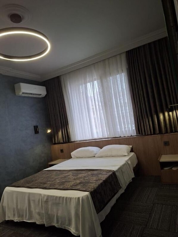 A hotel room with a bed, a bedside table, a wall-mounted air conditioner, and a window with curtains.