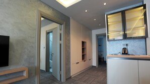 A modern hotel room with a flat-screen TV, a wooden cabinet, a built-in kitchen area, and a hallway leading to a bathroom. - Kaçkar Otel (Rize)