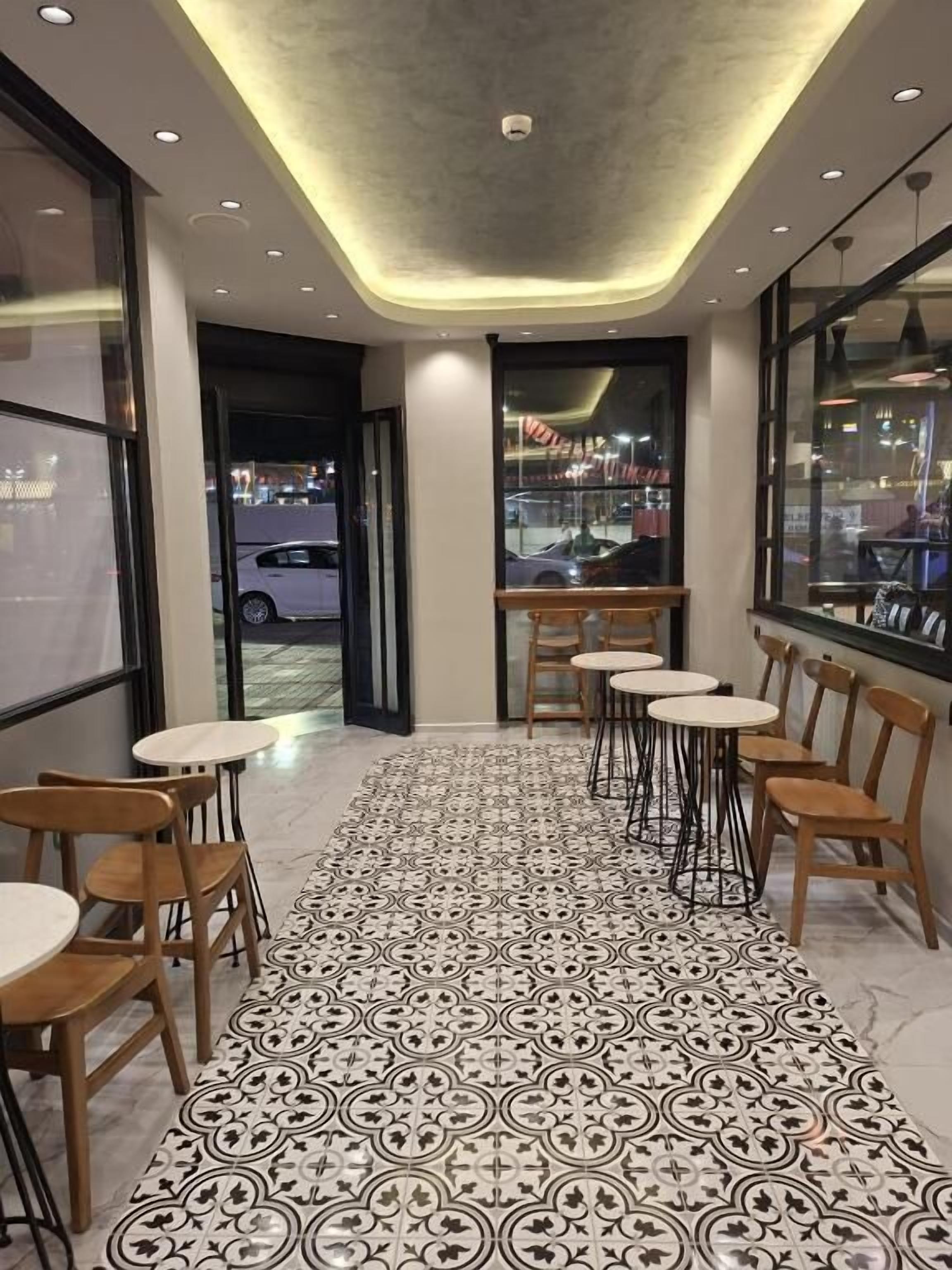 A corridor with patterned flooring, wooden chairs, and tables, leading to a window with a view of the outside.