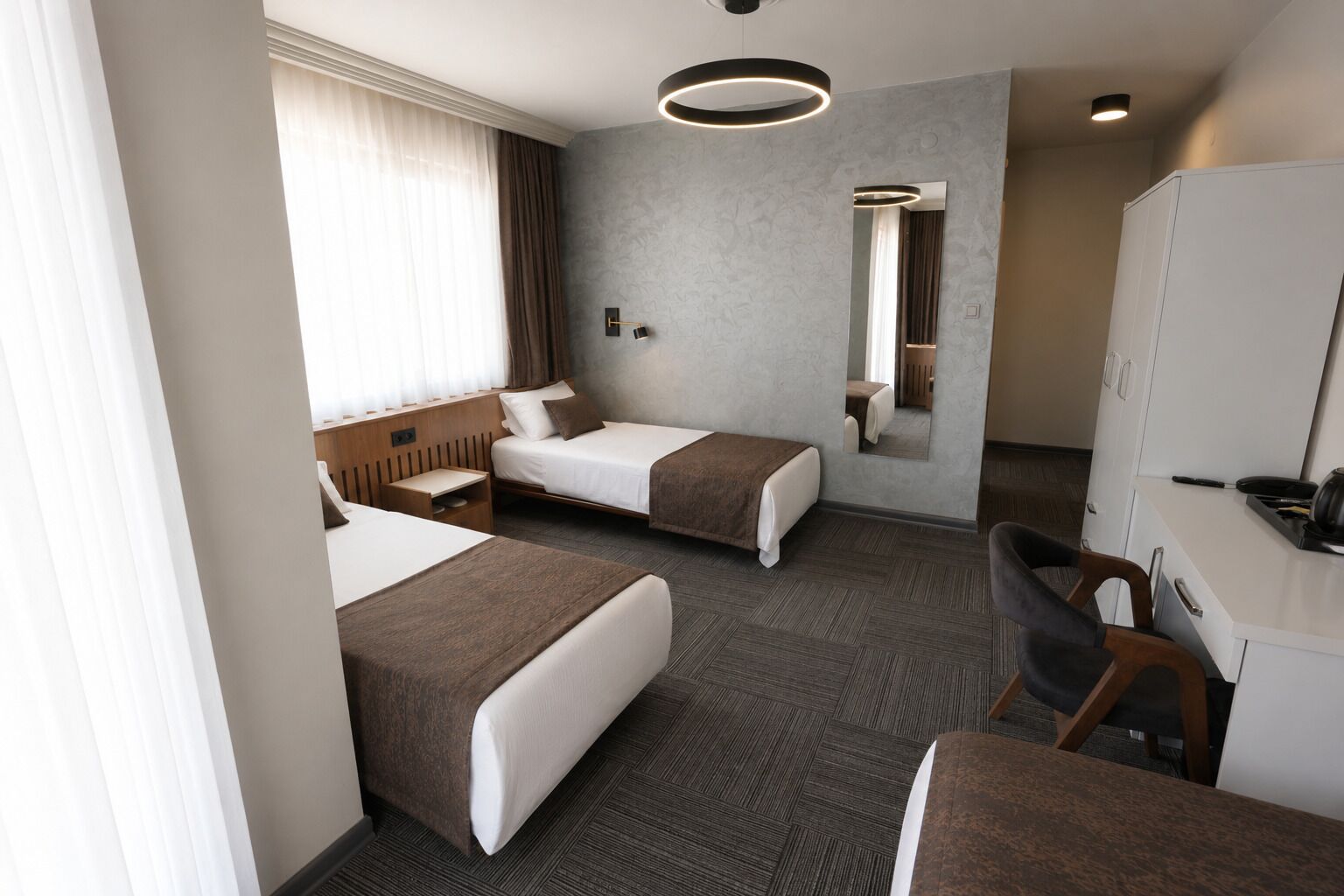 Quarto de hotel com duas camas, uma escrivaninha, uma cadeira, um espelho e luminária no teto.