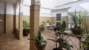 Un patio con suelo de baldosas, plantas en macetas, una mesa y sillas blancas, y arcos.