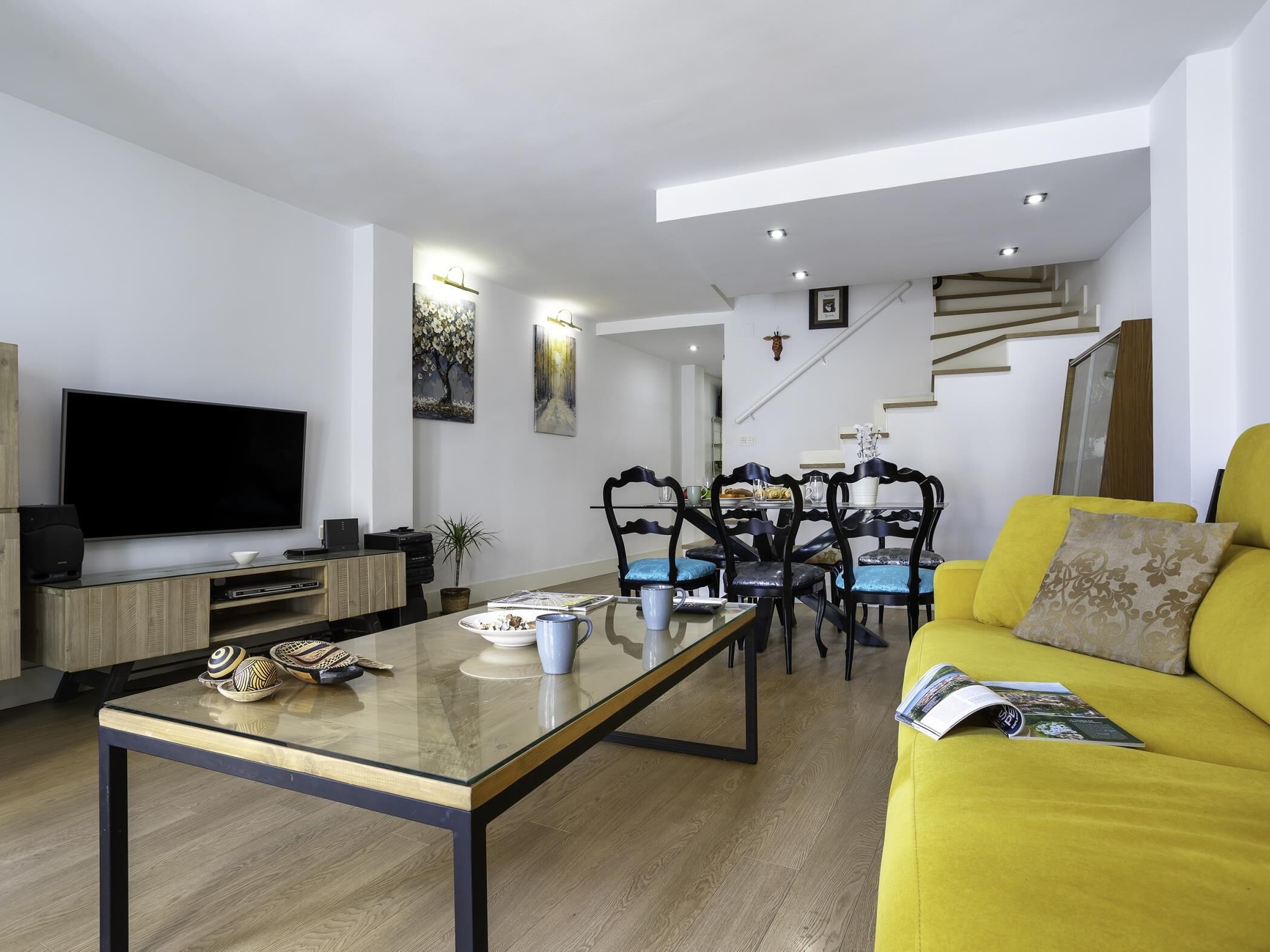 A modern living room with a flat-screen TV, a glass coffee table, a yellow sofa, and a dining area with chairs and a table.