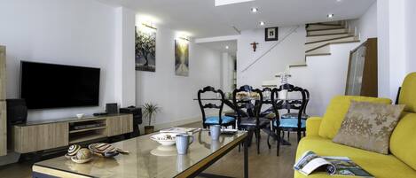 A modern living room with a flat-screen TV, a glass coffee table, a yellow sofa, and a dining area with chairs and a table.