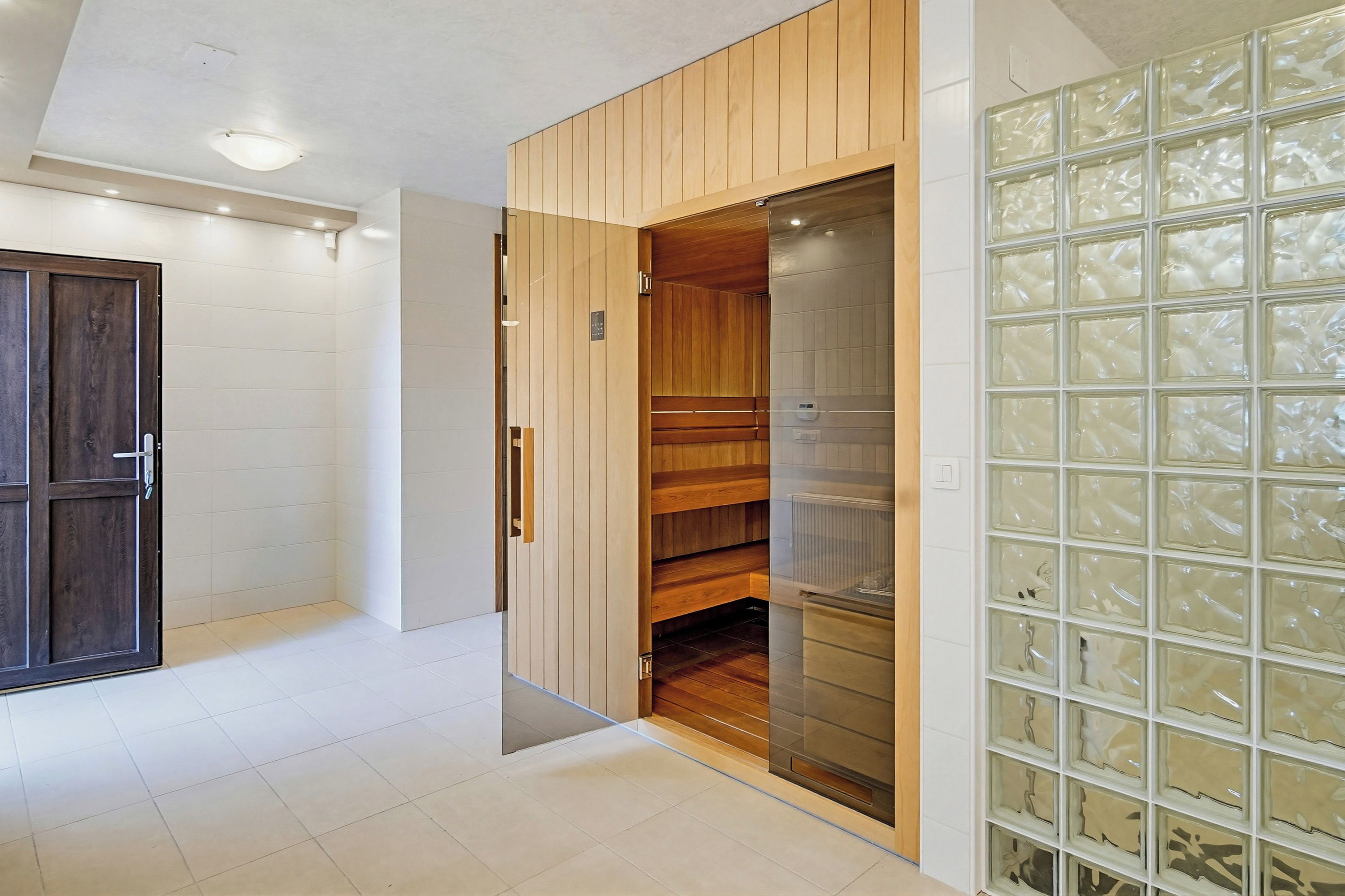 A room with a wooden door, a sliding glass door leading to a sauna, and a tiled wall with a geometric pattern.