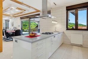 A modern kitchen with a central island, white cabinetry, a built-in stove, and a view of greenery outside. - Hillside Retreat With Pool & Wellness (Gornja Stubica)