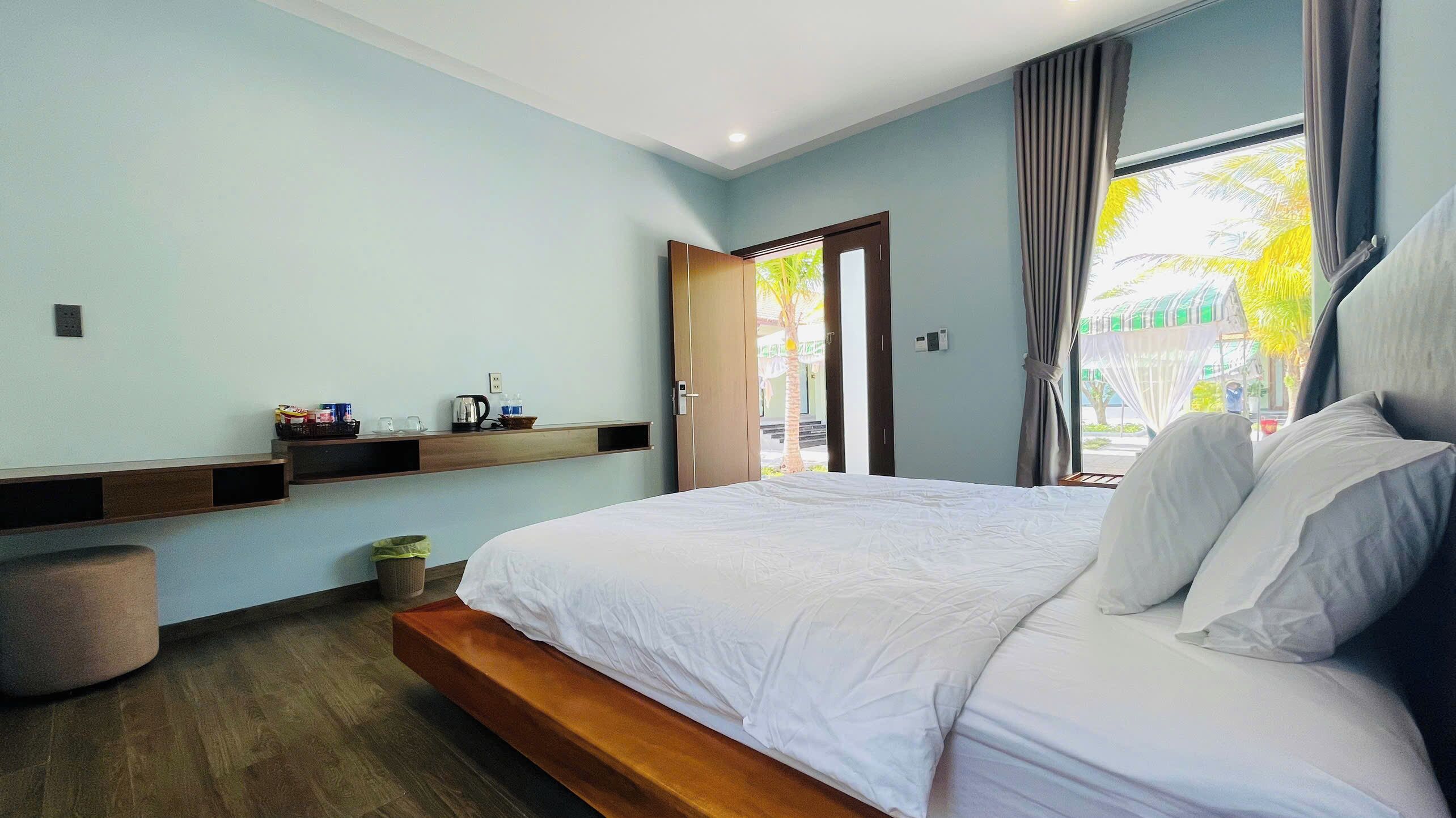 A hotel room with a bed, a small table with items, a trash bin, and a window with curtains.