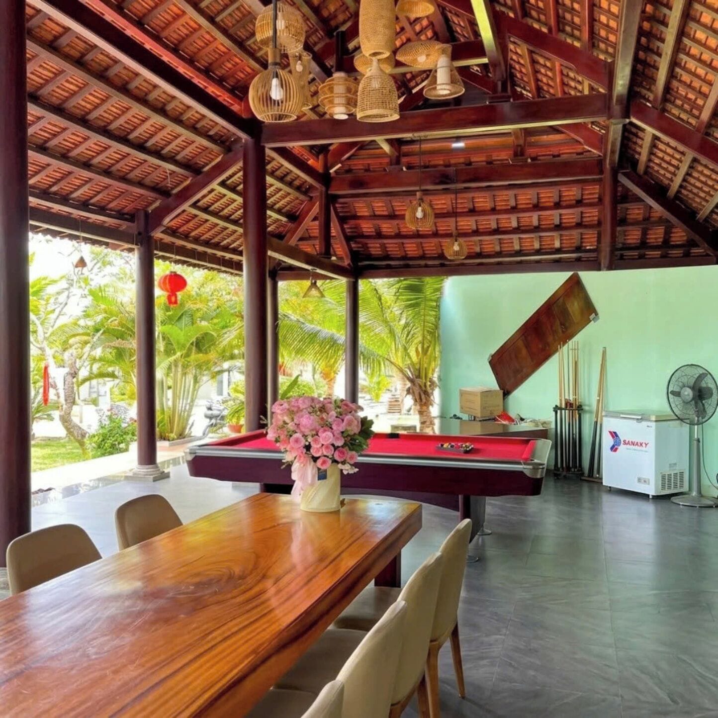 A pool table with a vase of flowers, wooden chairs, a ceiling with woven light fixtures, and a fan in a room with a green wall and a view of palm trees.