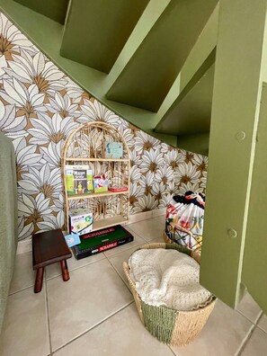 A room with a patterned wall, a small wooden shelf with items, a Scrabble game, a wicker basket with a cushion, and a door. - AppartCosy 20 Country House Near “Régalante” (Landavran)