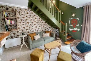 A living room with a staircase, a sofa, a coffee table, a wicker chair, a bookshelf, and a wall with a location pin graphic. - AppartCosy 20 Country House Near “Régalante” (Landavran)