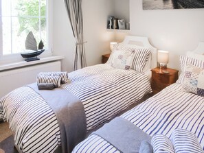 Une chambre avec deux lits, une fenêtre avec des rideaux, une table de chevet avec une lampe et une étagère avec des livres et un tableau encadré.