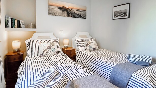 Een tweepersoonsbed met gestreept beddengoed, een nachtkastje met een lamp, een wandplank met boeken, en een ingelijste foto.