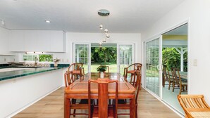 Una cocina moderna con armarios blancos, una mesa de comedor de madera con sillas y vistas a un jardín exuberante a través de puertas de cristal.