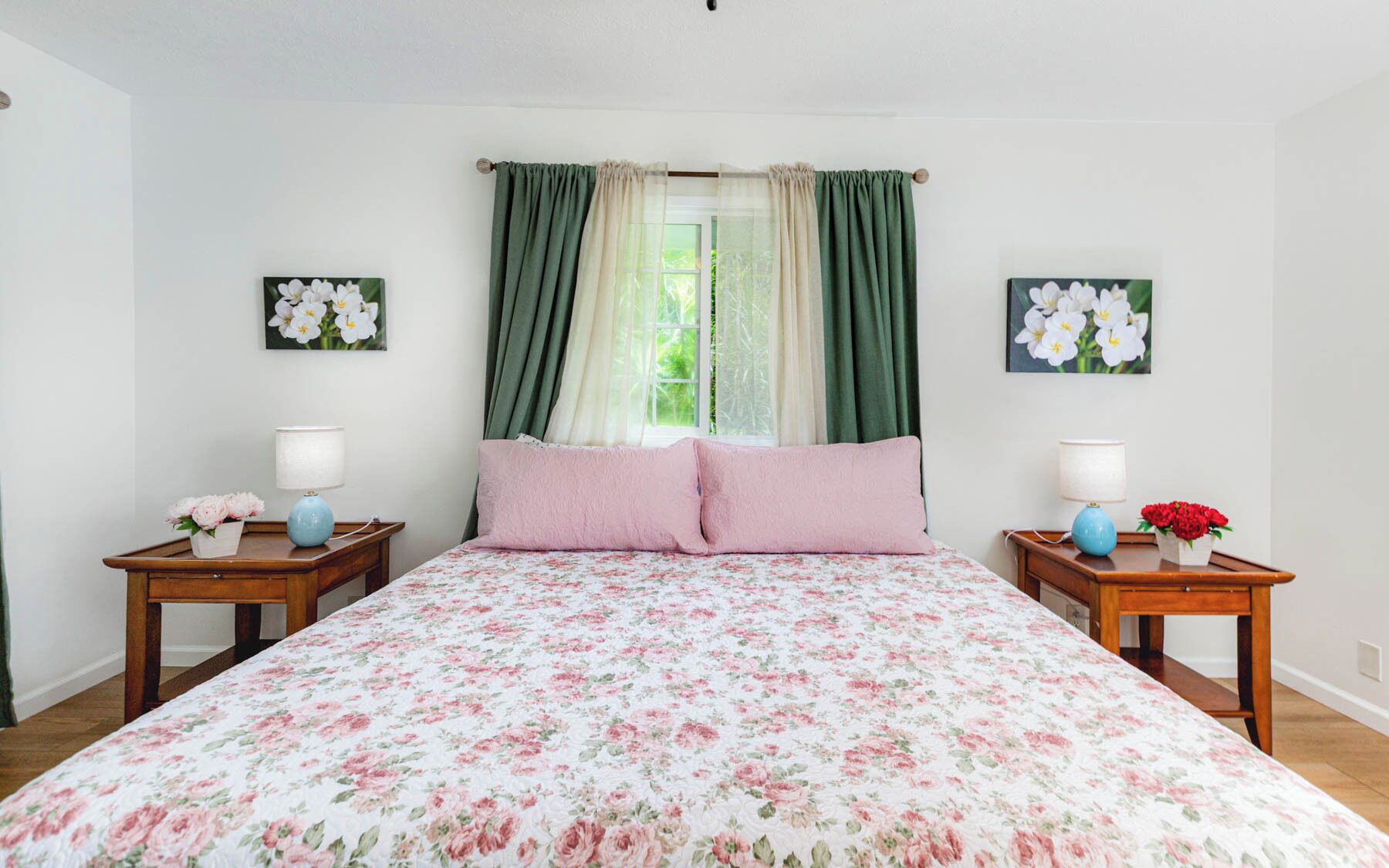 Un dormitorio con edredón estampado con flores, dos mesitas de noche con lámparas, una ventana con cortinas y decoración floral en la pared.