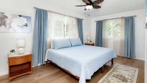 Un dormitorio con cama, dos mesitas de noche, ventilador de techo y ventana con cortinas.