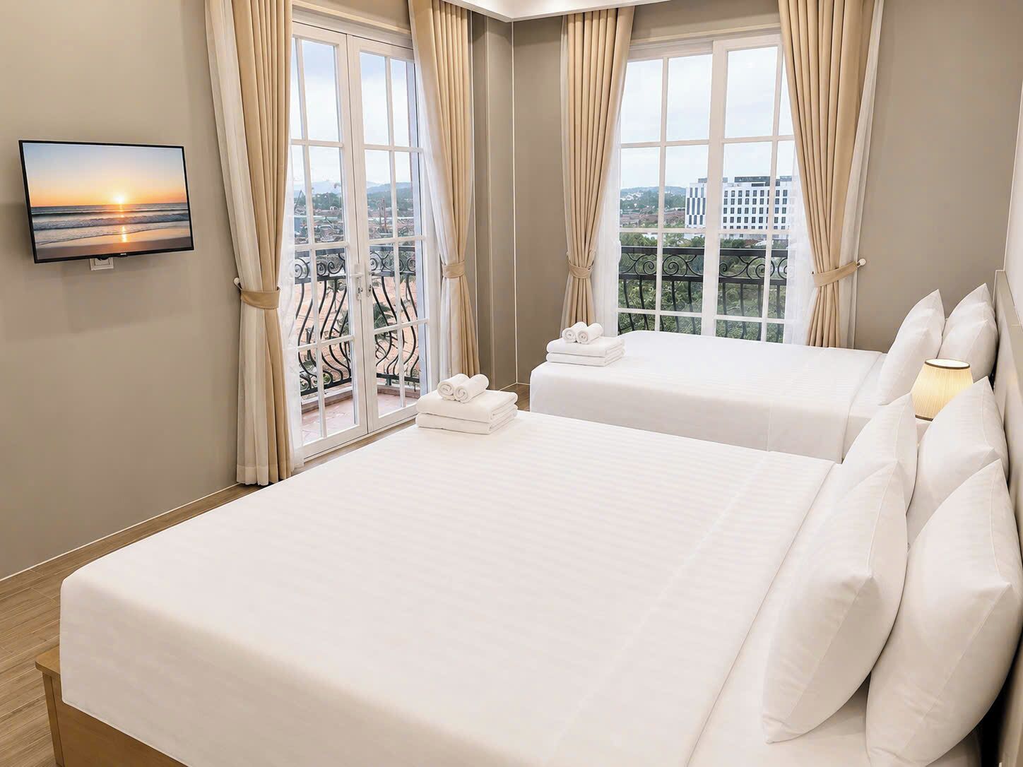 A hotel room with two beds, a TV on the wall, and a balcony with a view of the city.