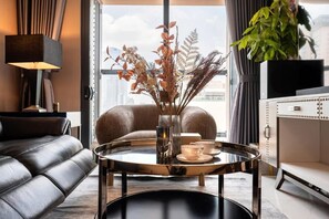 A modern living room with a leather sofa, a round coffee table with a vase and cups, a lamp, and a view of the city through large windows. - Madison modern 2br unit (Ho Chi Minh City)