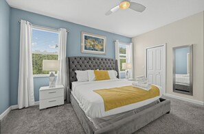 A bedroom with a tufted headboard, a bed with white bedding and a mustard throw, a nightstand with a lamp, a ceiling fan, and a window with blinds. - Game Room + Theater + Pool + Hot Tub | 15-Min Disney | Luxury 8BR Disney Villa (Davenport)