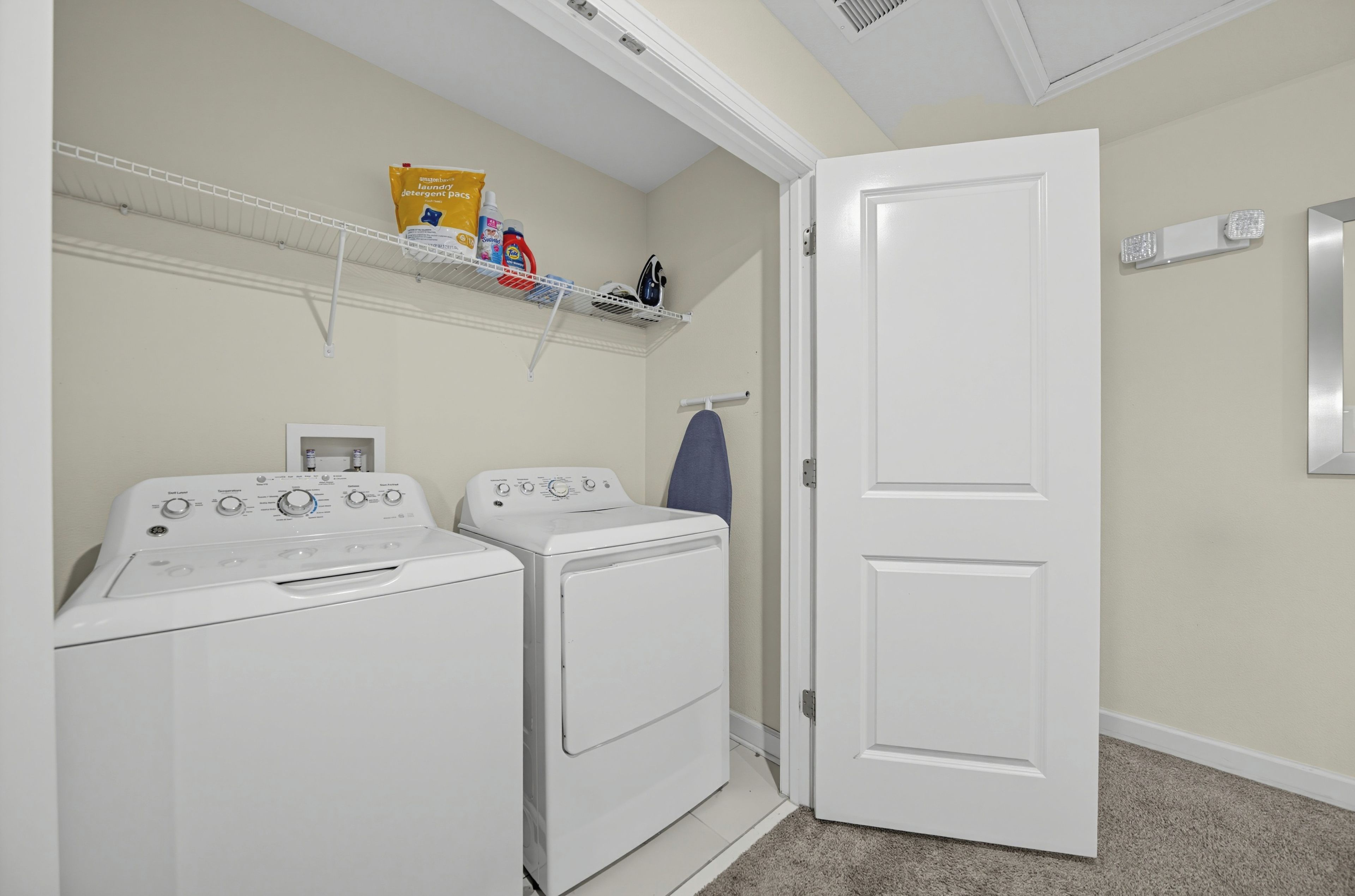 A laundry room with two washing machines, a shelf with detergent, and a door.