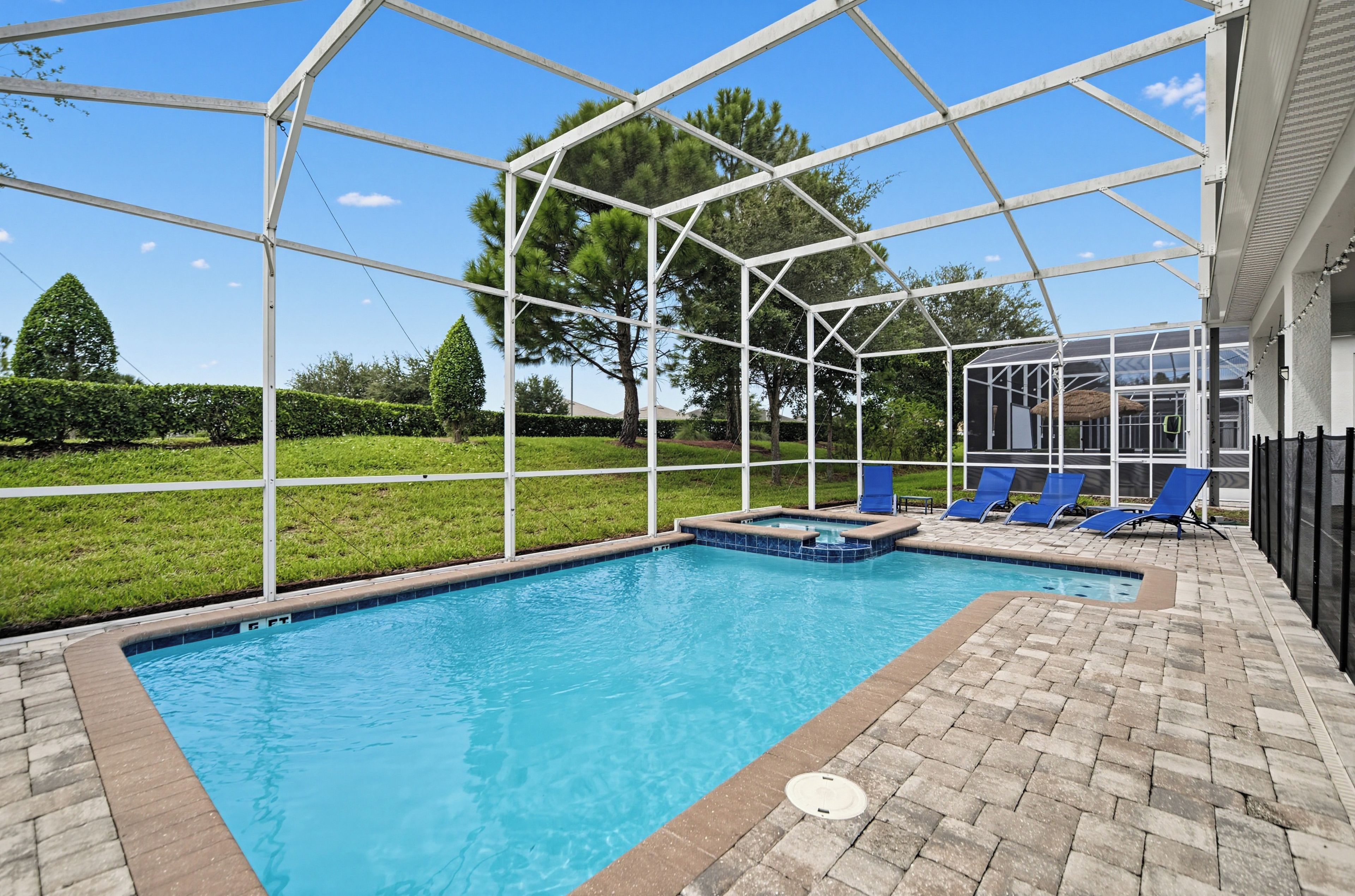 An outdoor swimming pool with a clear blue water, surrounded by a white metal structure, and a brick patio with blue lounge chairs.