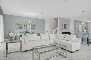 A modern living room with a white sectional sofa, a glass coffee table, and a dining area with blue chairs. - Game Room + Theater + Pool + Hot Tub | 15-Min Disney | Luxury 8BR Disney Villa (Davenport)